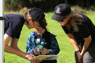 2 young women with WWT logos helping a young girl to identify an insect by Lauren Roberts