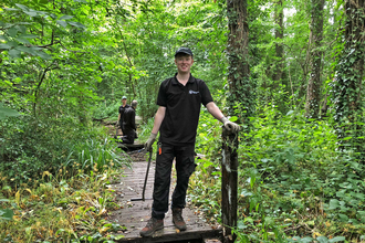 Man in a Worcestershire Wildlife Trust logo'd polo shirt standing on the end of a boardwalk in a woodland, looking at the camera and smiling