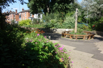 A circular bench in the middle of a flower-rich greenspace with houses in the background by Liz Yorke