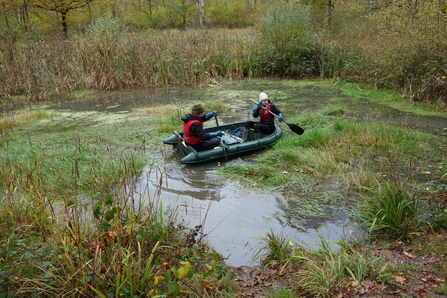 Two people in a boat on a pond, rowing to reach the Crassula that needs removing by Lydia Rackham