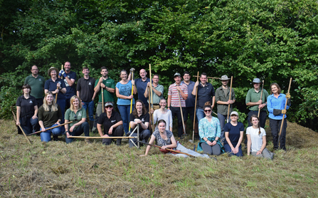 Group of Worcestershire Wildlife Trust, many holding tools to rake a meadow, in a meadow by Jess Nott