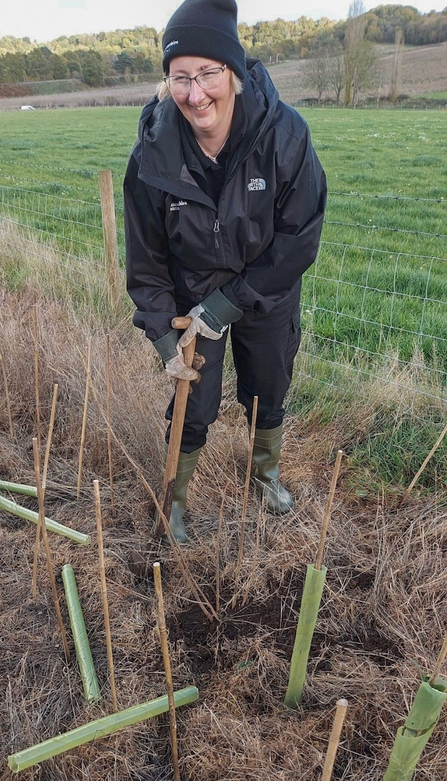 Woman in waterproofs and a woollen hat digging a hole to plant a hedgerow