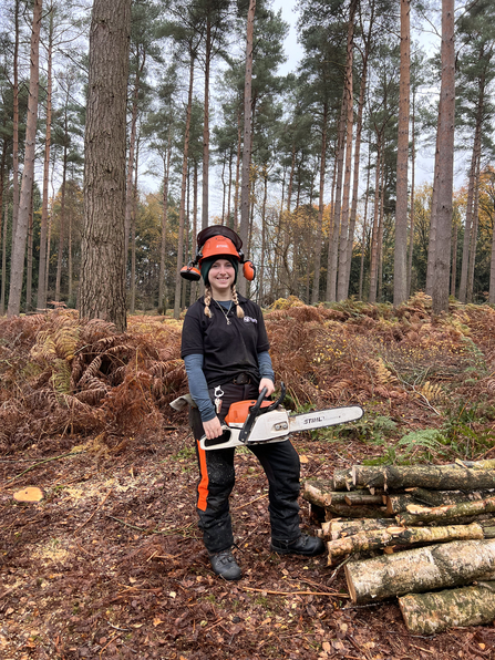 Woman in protective wear clothing holding a chainsaw by a pile of logs