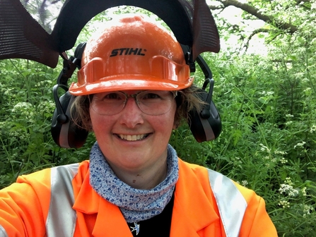 Woman wearing a chainsaw helmet with visor up is in a woodland and is smiling at the camera