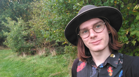 Person in a hat and glasses with a black shirt with a pattern of brightly coloured feathers looking at the camera. They're in front of a mature hedge.