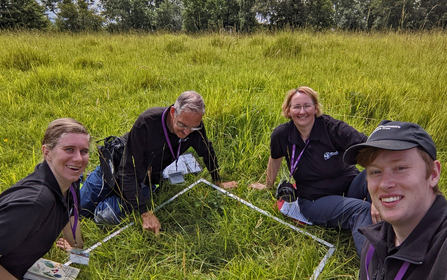 Four trainees sitting around a quadrat (surveying square) in a meadow - all are enjoying themselves, three are smiling at the camera
