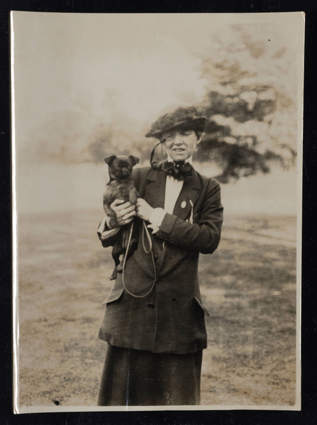 Sepia-toned photo of a woman in Edwardian dress, with a hat and monocle, in a field with a tree behind her and holding a dog