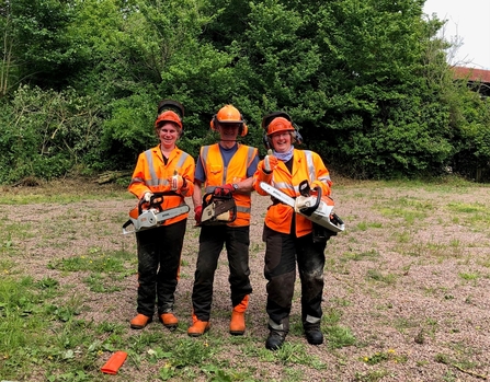 Three people in high-vis safety clothing holding chainsaws - the two women on the outside of the trio are holding up their thumbs after passing their chainsaw assessement