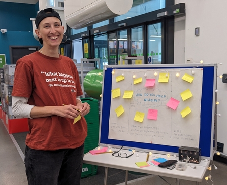 Person in an burnt orange t-shirt standing near doors in a large shop and next to a board with some paper and post-it notes on