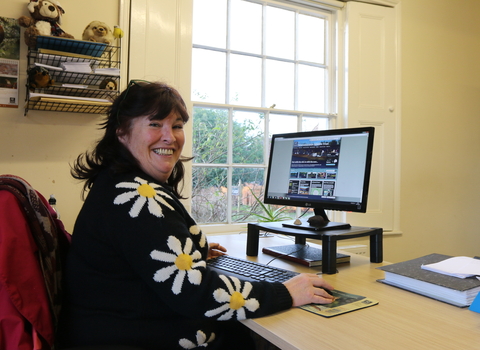 Woman in a top with large daisy flower pattern sitting at a computer but looking at the camera but w