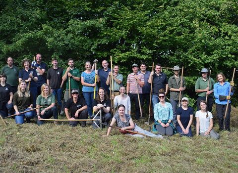 Group of Worcestershire Wildlife Trust, many holding tools to rake a meadow, in a meadow by Jess Nott