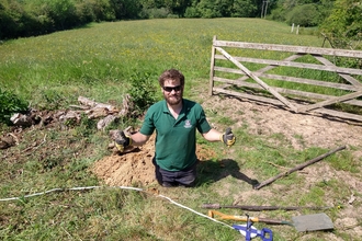 Young man in the middle of a job in a field on a sunny day who's paused and is giving 'thumbs up' to the camera