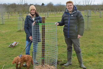 A woman (in woolly hat) and man (in insulated jacket) standing on either side of a recently planted tree