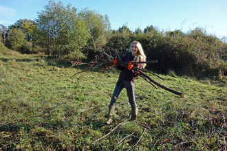 Woman with blond hair, laughing, carrying a pile of brash (sawn-off tree branches) by Iain Turbin