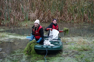 Two women in a small rowing boat removing green vegetation from a pond by Iain Turbin