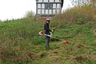 Woman wearing a helmet and holding a brush-cutter on a grassy bank by Iain Turbin