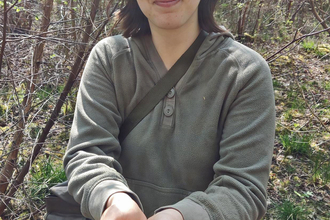 Woman with dark hair, smiling and looking at the camera as she holds a dormouse in the palm of her hands