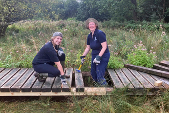 Two women in/on a boardwalk, repairing it, both looking at the camera and smiling