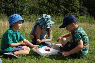 Three children with minibeast pots in a grassy field by Lauren Roberts