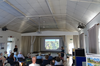 A village hall with a woman at the front pointing to a screen with a map on and a few rows of seated people (by Julie Grainger)