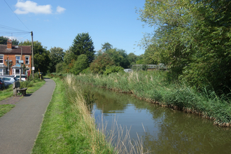 Canal on the right hand side with a path running away from the viewer on the left, flanked by parked cars and terraced houses by Liz Yorke