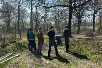 5 people standing in a woodland as they watch a 6th person demonstrate how to use a petrol auger