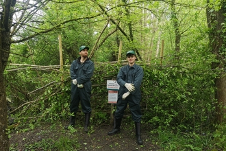 Two people standing in front of a dead hedge and signs