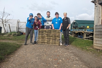 A group of people standing behind a hurdle that they've woven - three people on the left are grinning and pointing at the camera/holding their thumbs up