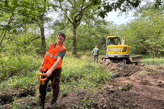 A woman in a high-vis jacket is smiling at the camera - she's standing in a woodland in summer and there's a recently dug trench behind her
