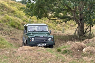 A Land Rover in a lumpy off-road situation; there's a hill to the left, a tree to the right with a hump in front of the vehicle