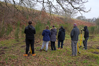A group of people standing and looking at vegetation along the side of a railway line. It's a misty day in winter and everyone is wearing coats, some people are wearing hats.