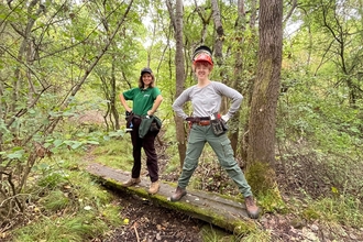 Two women standing on a plank and posing for the camera. They're in a woodland and are both smiling.