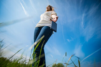 A woman walking through a field on a sunny day; she's dropping her phone