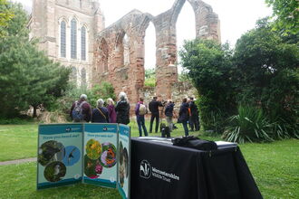 A group of people standing between a display of Worcestershire Wildlife Trust information and the ruins of the abbey at Worcester Cathedral