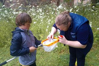 An adult with long hair tied back and a gilet is helping a child look into a net - they're also holding a bug pot
