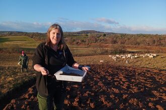 A woman with long hair smiling at the camera and holding a tray of seed that she's spreading by hand; it's a sunny day and she's standing in a field with sheep in the distance