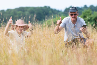 Two volunteers are crouching/sitting in amongst vegetation on a summer day and both are looking at the camera with their thumbs up