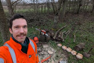 A man with dark hair and a moustache is wearing a high vis jacket and is taking a selfie - in the background you can see some felled wood and a chainsaw