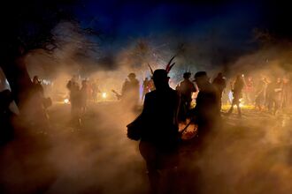 People are silhouetted by backlight on an evening in an orchard - the person closest to the camera has feathers in their hat