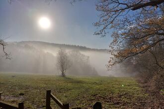 A photo of a landscape with a field in the foreground and a line of hills in the background. The sun is shining and the sky is blue and there is a blanket of mist covering the hills.