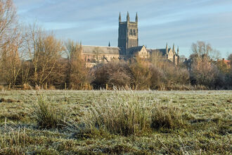 A large cathedral with a square tower is in the distance and we're looking at it over a piece of rough grassland with a hedge in the mid-distance. It's a sunny day but frost is on the ground and there are no leaves on trees.