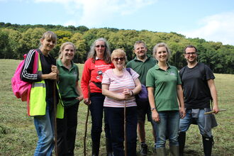 A group of people smiling at the camera - they're in a meadow on a sunny day and are holding tools