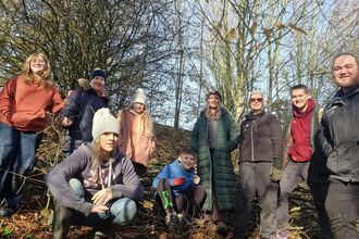 A photo of nine people smiling at the camera, some are standing and some are crouching. They are outdoors in a woodland with trees behind.