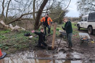 A photo of three trainees smiling at the camera standing and sitting around a post that they are installing in the muddy ground next to a big puddle of water.