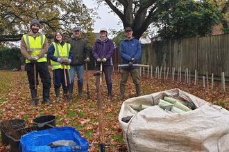 A group of five people holding spades and smiling at the camera standing next to trees that have just been planted with a fence behind and orange leaves on the ground
