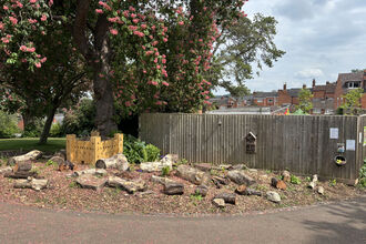 A photo of a bug hotel in a park with lots of logs and a mini castle with a fence a tree with pink flowers behind