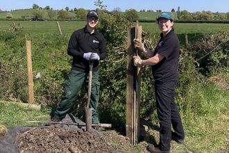A photo of two conservation trainees standing at the edge of a field smiling at the camera whilst installing a post in the ground