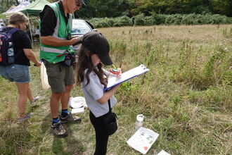 A photo of a girl standing in a meadow holding a clipboard doing bug hunting with two adults standing behind
