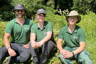 A photo of three trainees wearing hats and green branded polo tops sitting on the ground and smiling at the camera