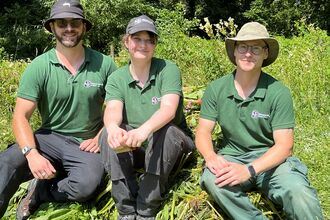 A photo of three trainees wearing hats and green branded polo tops sitting on the ground and smiling at the camera